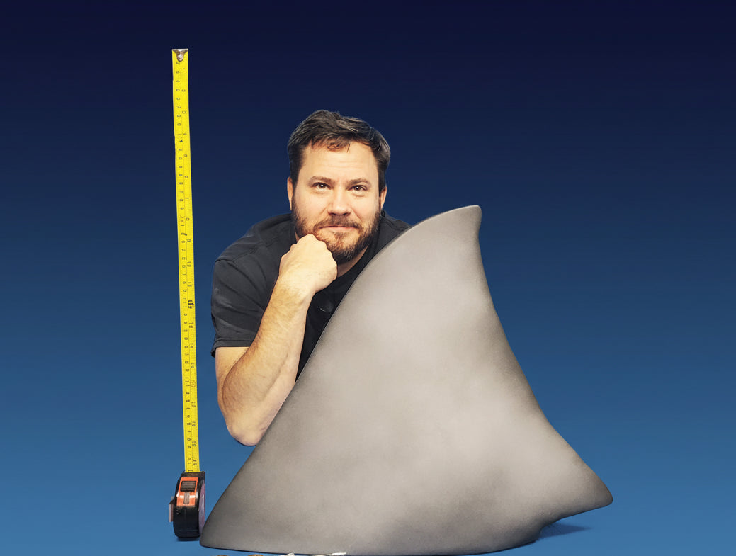 Man holding a large shark fin with a measuring tape against a blue background/ jaws shark fin